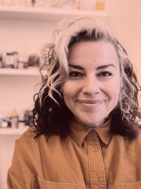 A photo of Alison Wren, a white person with long hair and nose piercings. They are smiling at the camera. They are wearing a yellow shirt buttoned up to the top and standing in front of some shelves of art materials.
